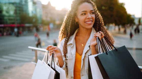 Imagem de uma mulher negra, de cabelos encaracolados e compridos, carregando vrias sacolas de compras para exemplificar o que  valor agregado.