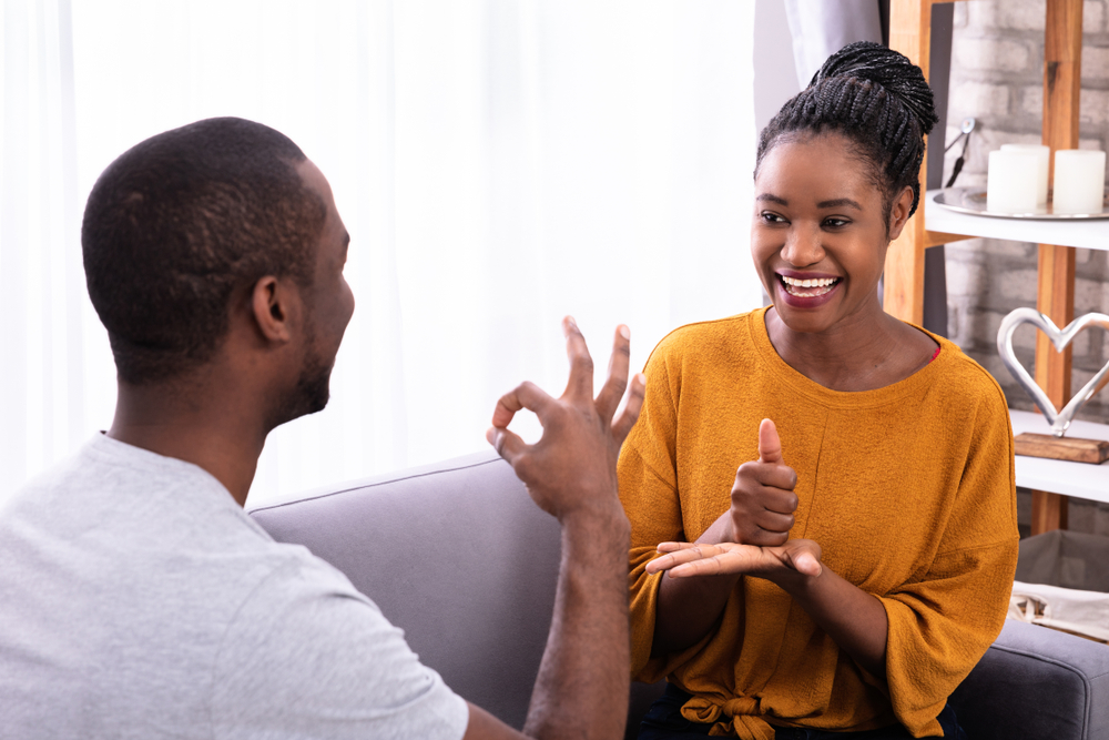 Um homem e uma mulher negros esto sentados em um sof cinza e conversando atravs de linguagem de sinais.