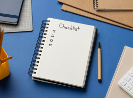 Caderno em cima de uma mesa azul com anotao de checklist mostrando o que  ser produtivo. Em volta do caderno, alguns itens de escritrio como calculadora, lpis e agendas.
