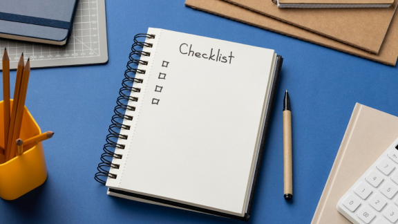 Caderno em cima de uma mesa azul com anotao de checklist mostrando o que  ser produtivo. Em volta do caderno, alguns itens de escritrio como calculadora, lpis e agendas.