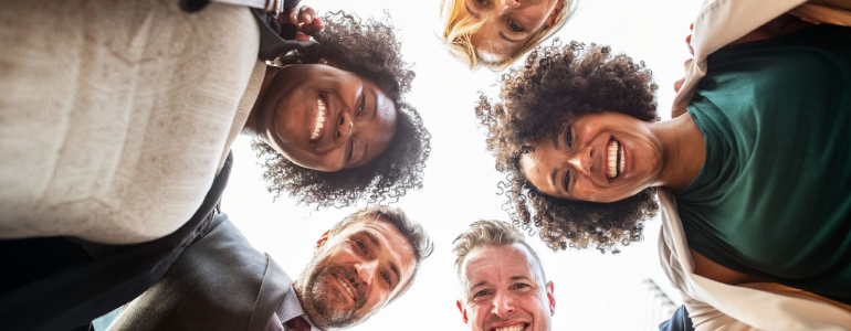 Grupo de pessoas reunidas em uma roda, esto sorrindo enquanto olham para baixo. A imagem representa habilidades socioemocionais em um ambiente de trabalho.