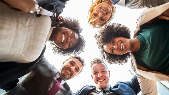 Grupo de pessoas reunidas em uma roda, esto sorrindo enquanto olham para baixo. A imagem representa habilidades socioemocionais em um ambiente de trabalho.