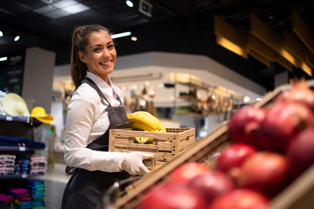 Uma mulher vestindo avental reabastecendo uma prateleira de frutas de um supermercado.