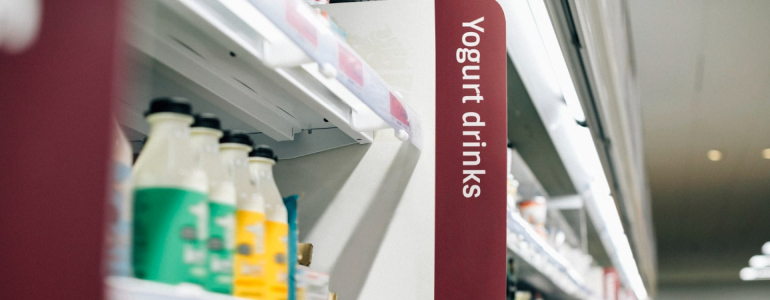 Prateleira de supermercado cheia de produtos. O elemento em destaque da imagem  a placa que indica a seo de iogurtes do estabelecimento, que traz a identidade visual do local.