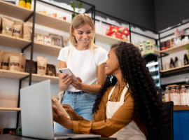Duas mulheres estão em uma loja, uma sorri enquanto segura a mercadoria em frente a um notebook, e a outra está com o celular em mãos. Ao fundo, existe um estoque cheio e com produtos variados, ilustrando os diferentes tipos de varejo.