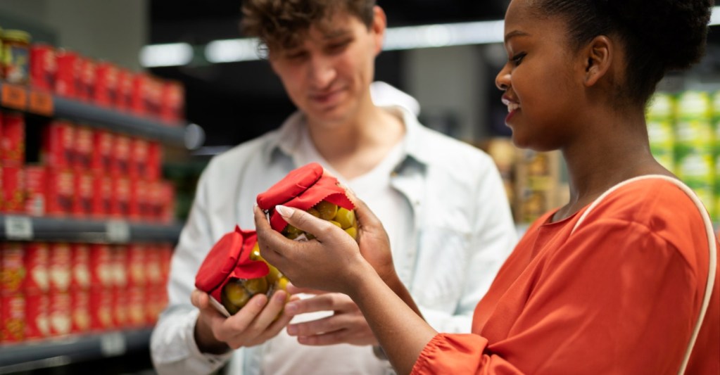 Uma mulher e um homem no supermercado escolhendo um produto para levar. Ambos esto com potes do mesmo produto em mos.