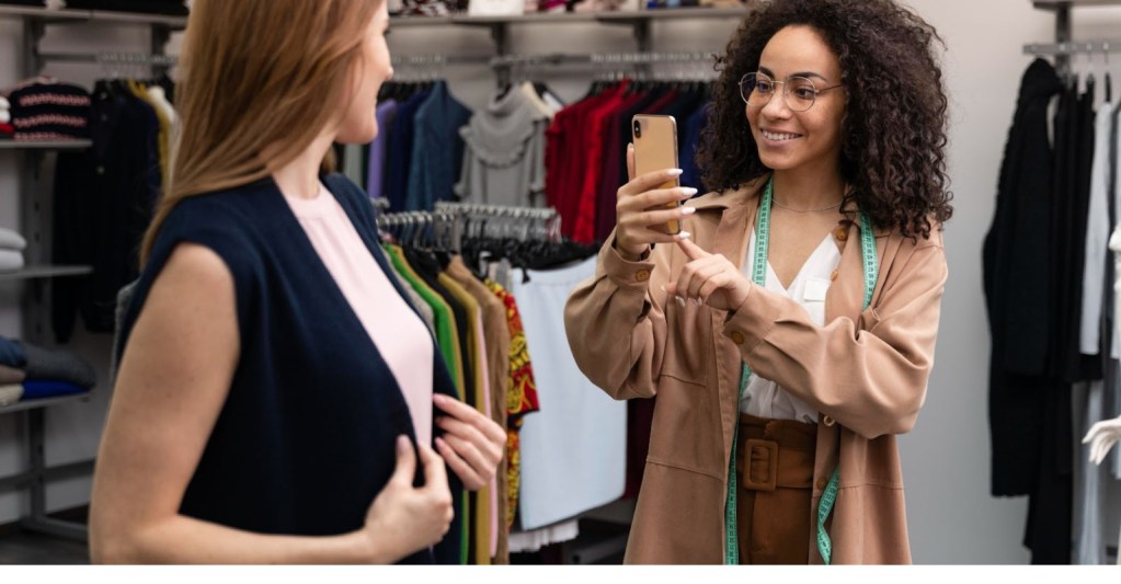 Duas mulheres em uma loja de roupas. Uma delas, que aparenta ser a vendedora, est tirando uma foto da cliente com uma pea de roupa.